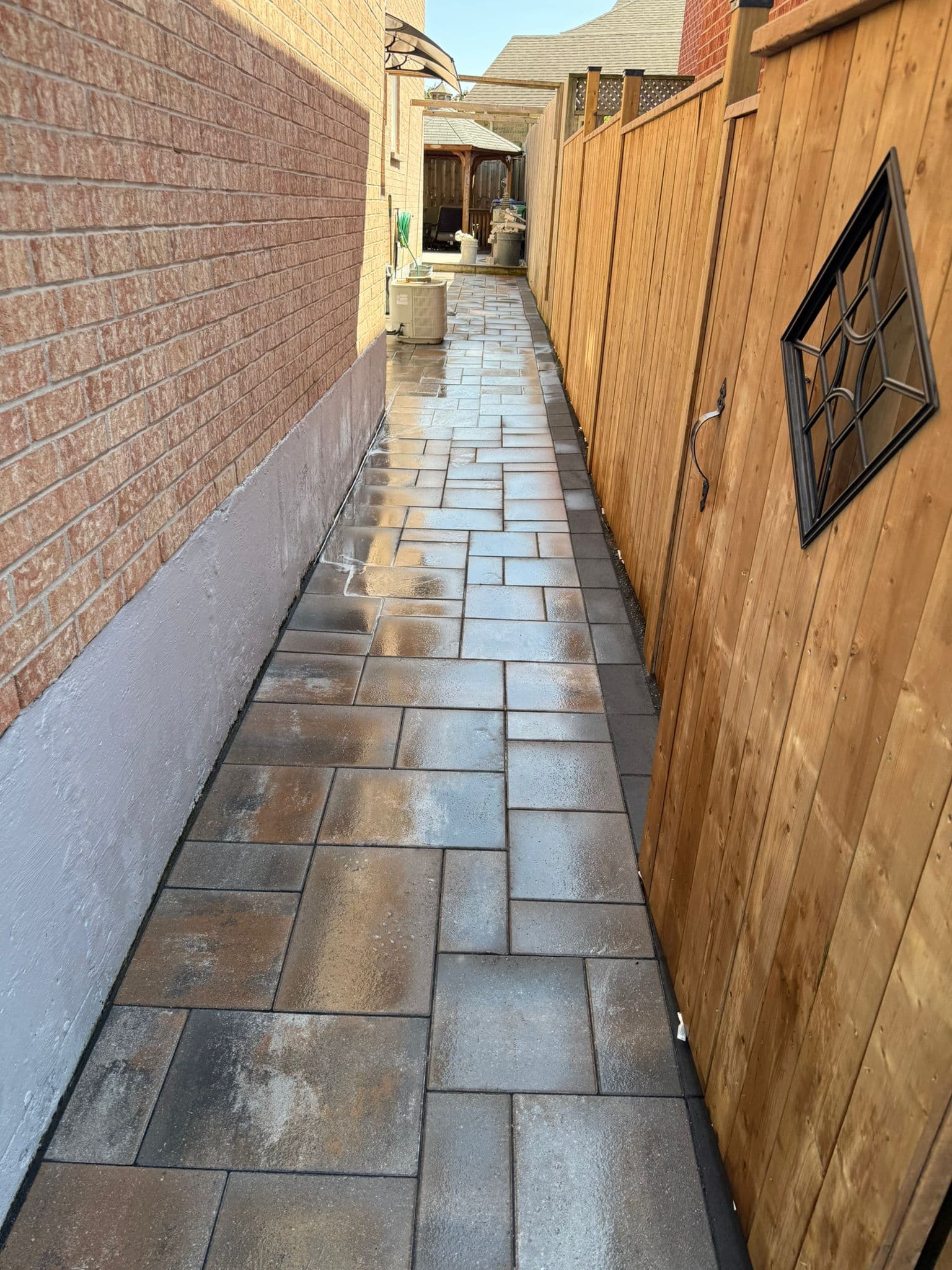 A narrow, wet stone paver walkway between a brick house and a wooden fence.