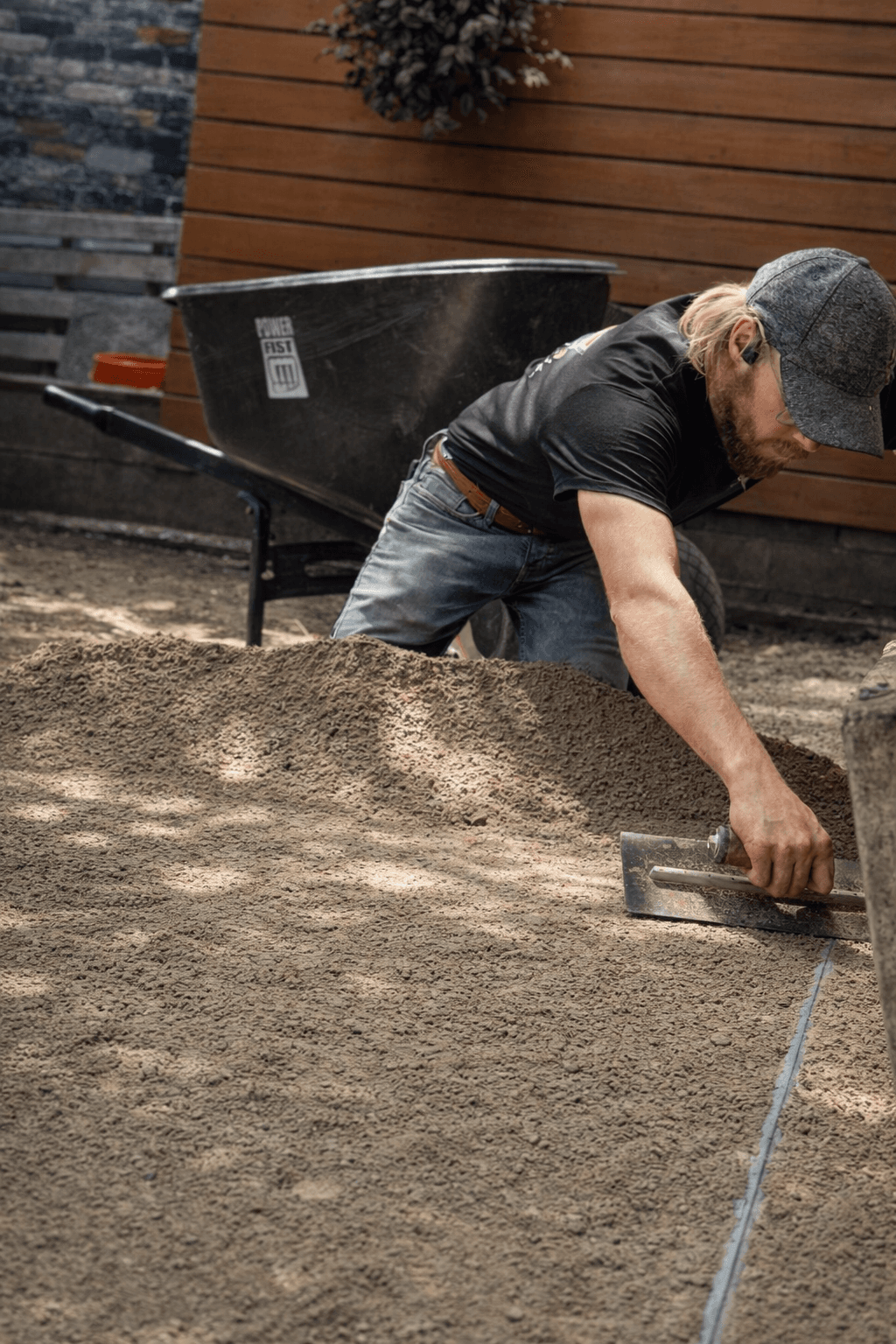 Bearded man in a cap levels sand with a trowel near a black wheelbarrow.
