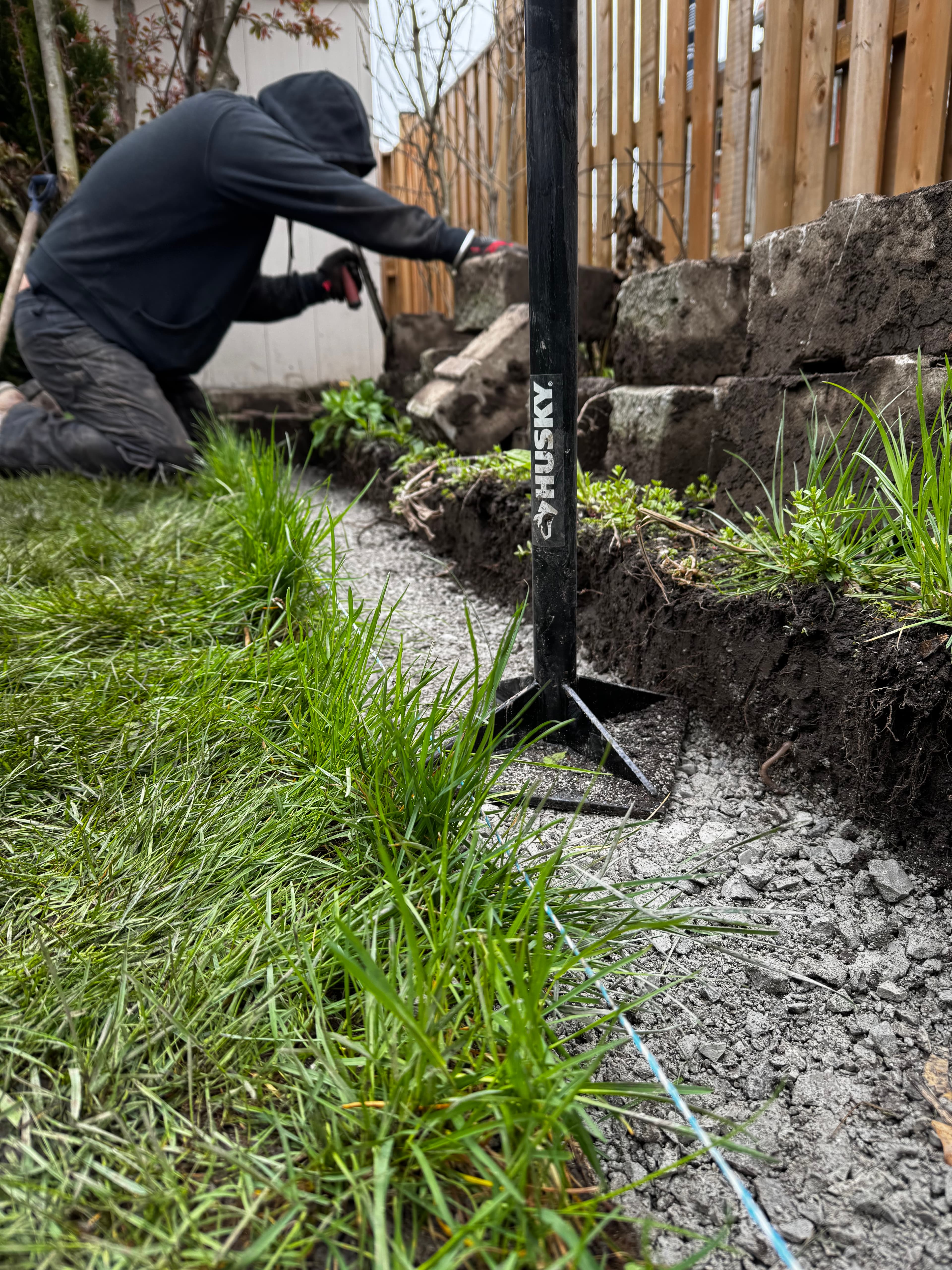 Person in a black hoodie uses a Husky tool to dig between grass and stone.