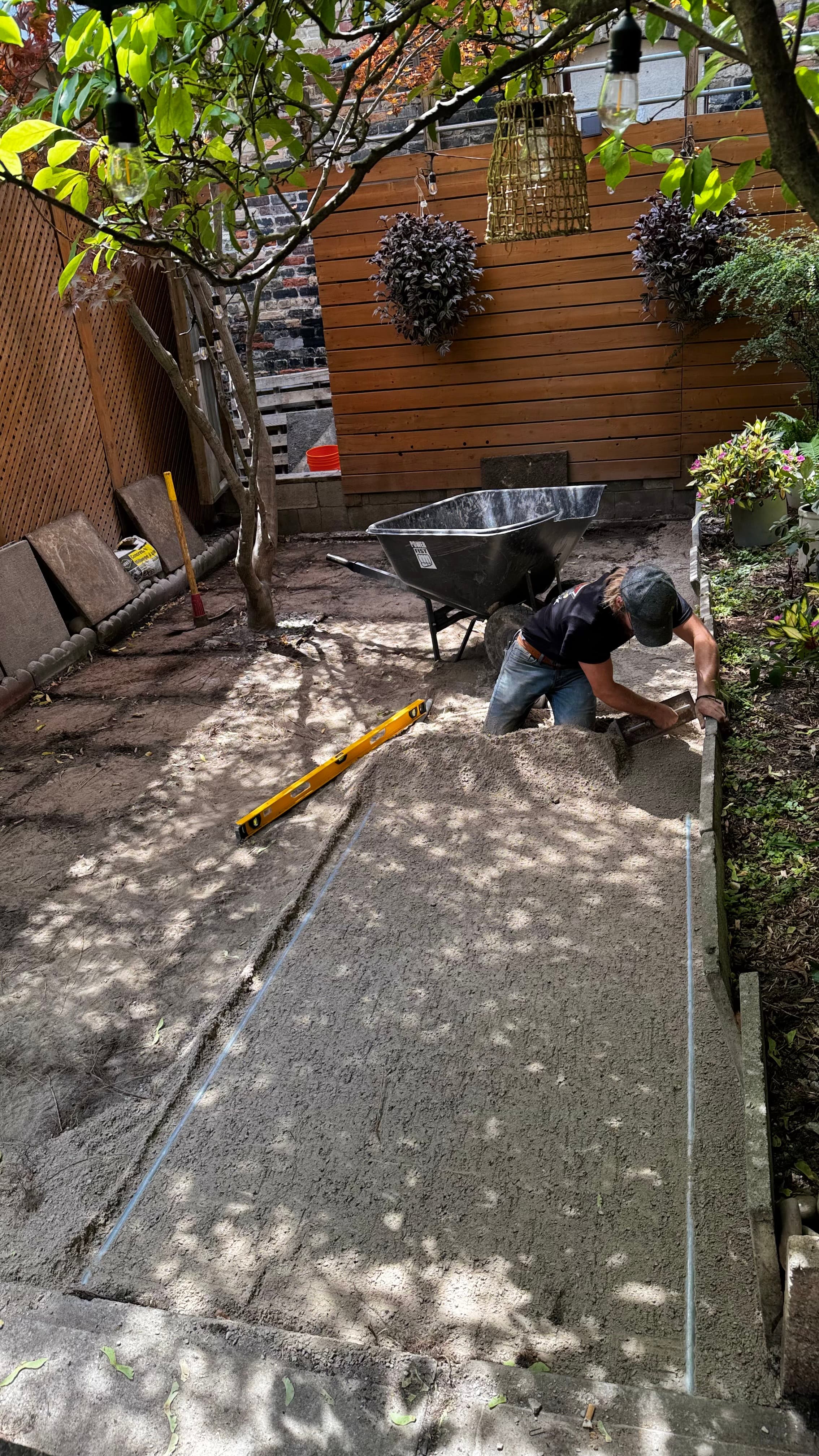 Man levels sand for a patio base in a backyard with a wheelbarrow and level.