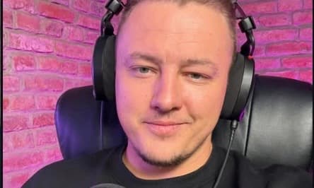 Man with a slight smile wearing black headphones sitting against a pink brick wall.