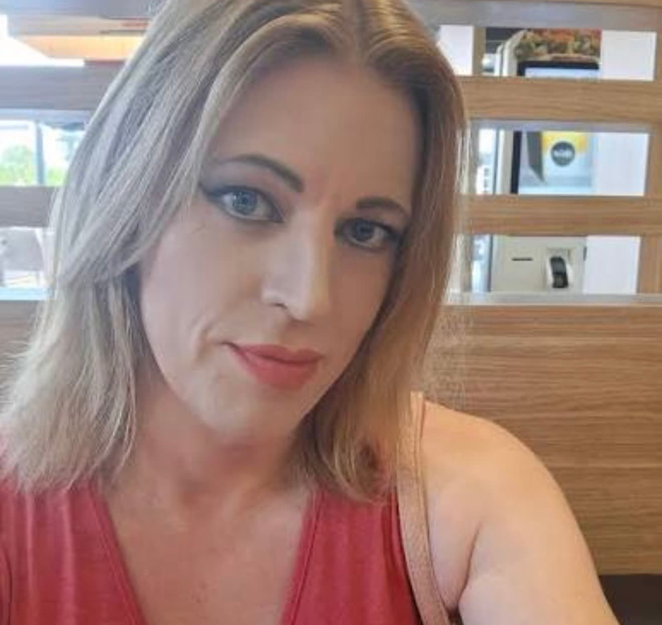 Blonde woman with blue eyes and makeup wearing a red top in a wooden booth.