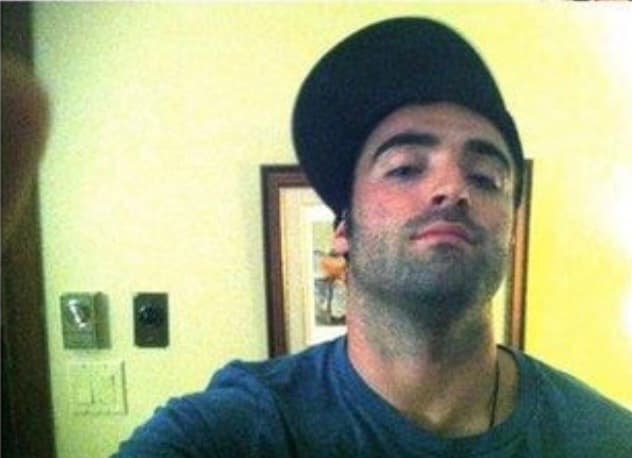 Man with stubble wearing a blue t-shirt and a tilted black baseball cap.