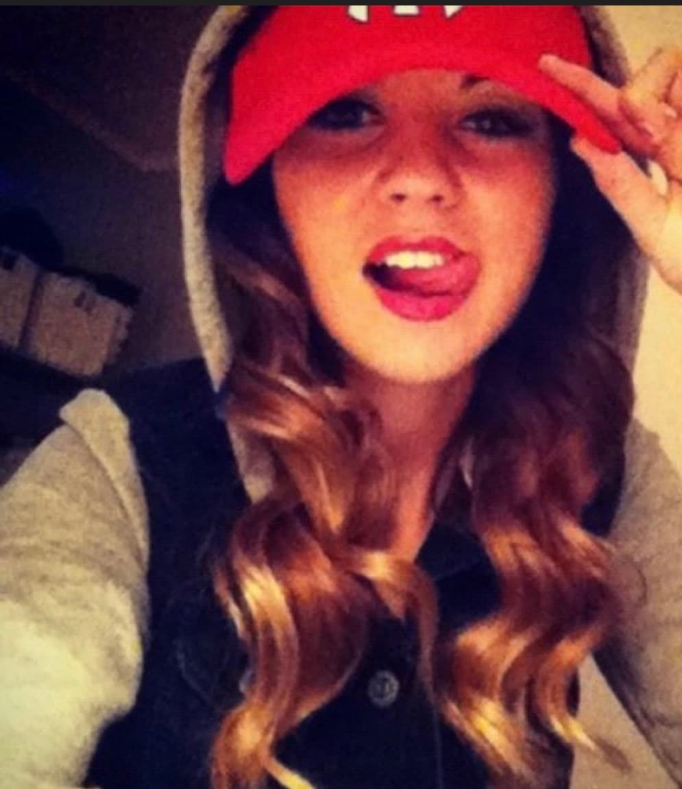 Young woman with wavy hair, red cap, and grey hoodie, sticking her tongue out playfully.
