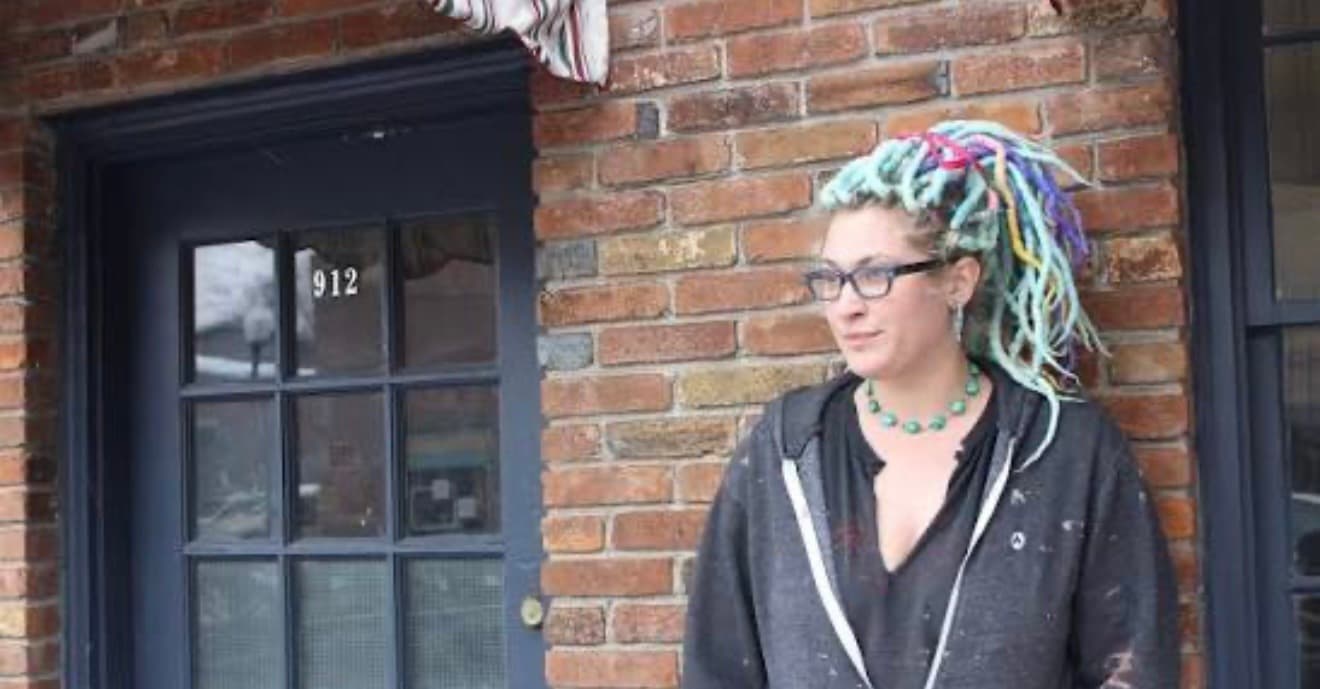 Woman with vibrant multi-colored dreadlocks and glasses standing outside a brick building with blue door.