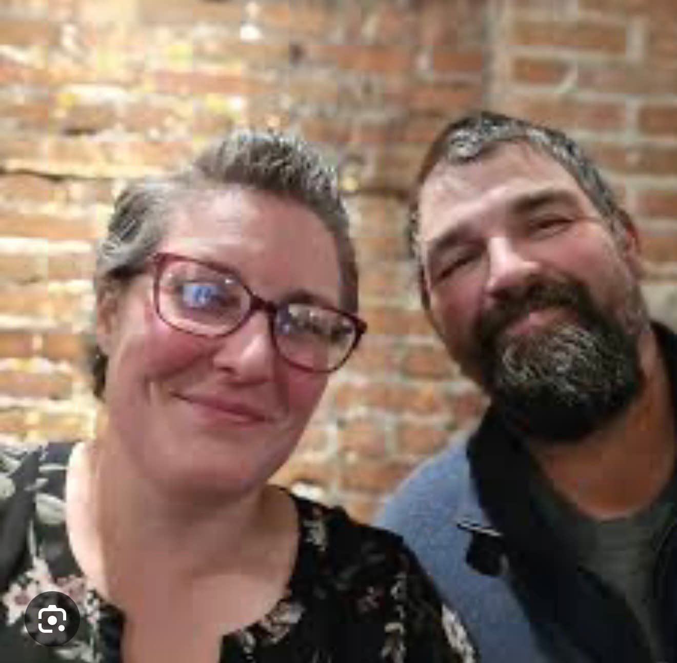Smiling woman with red glasses and a bearded man posing against a brick wall.