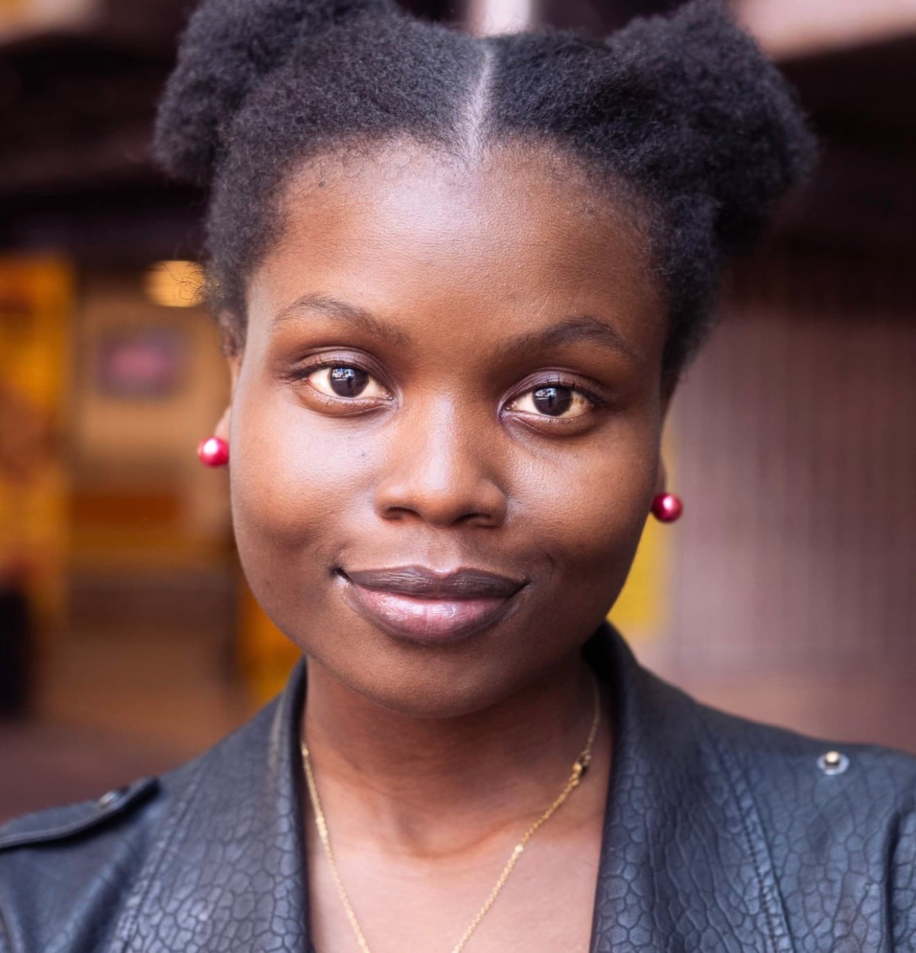 Smiling Black woman with afro puffs, red stud earrings, and a black leather jacket.